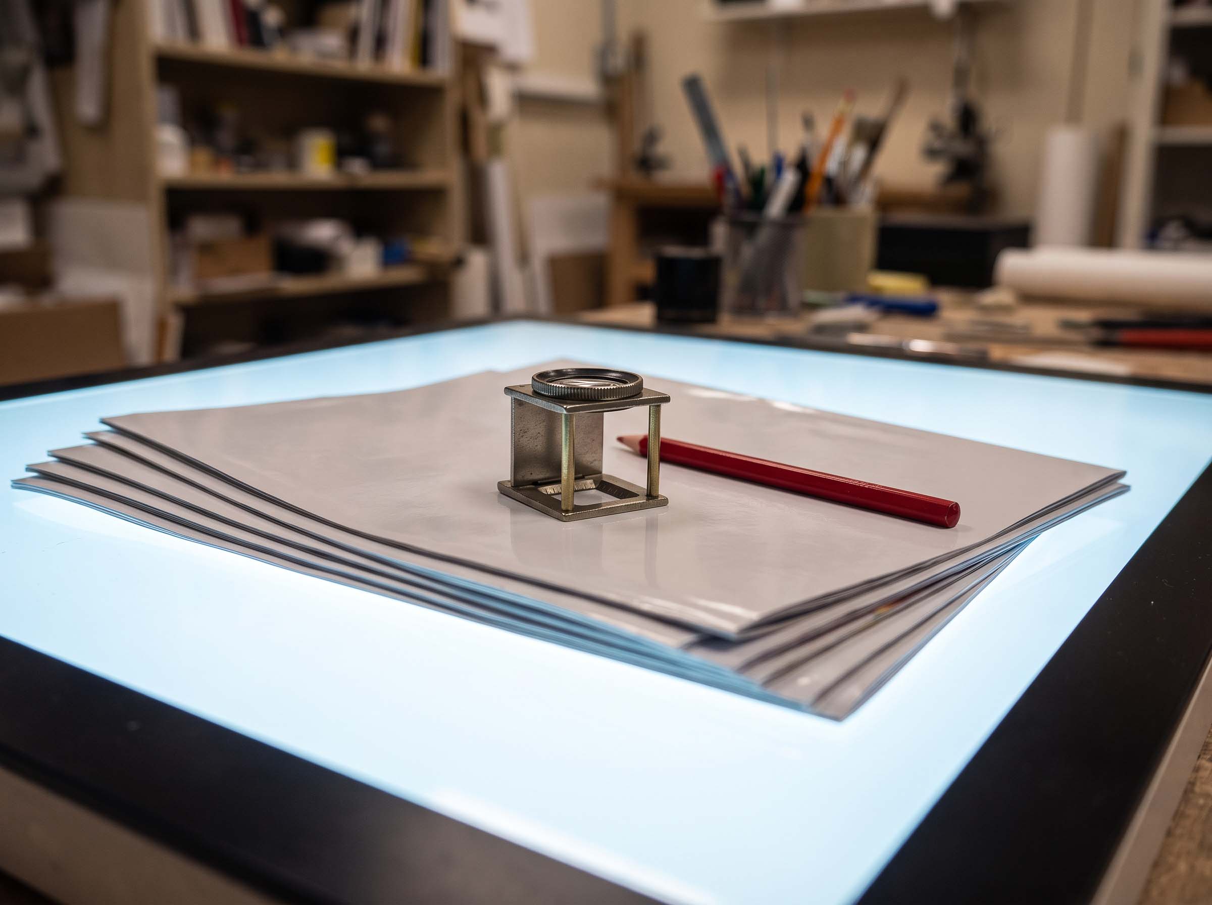 A retoucher's lightbox with magazine print proofs and a linen loupe resting on the sheets