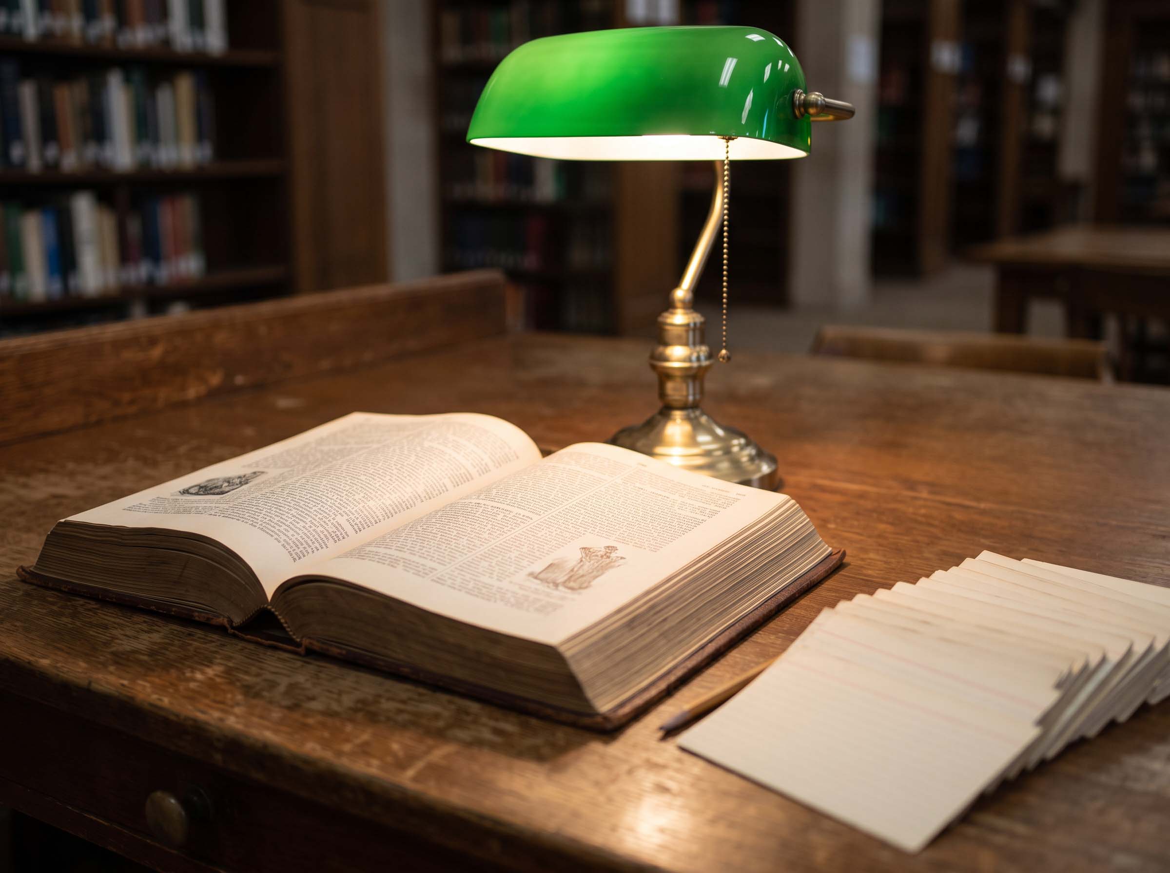 A heavy bound anatomical terminology volume open on a library reading desk under a green shaded lamp