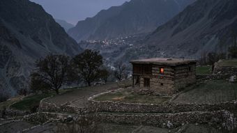 A lamplit stone seclusion house set apart below a terraced mountain village at dusk, illustrating menstruation taboos documented by anthropologists
