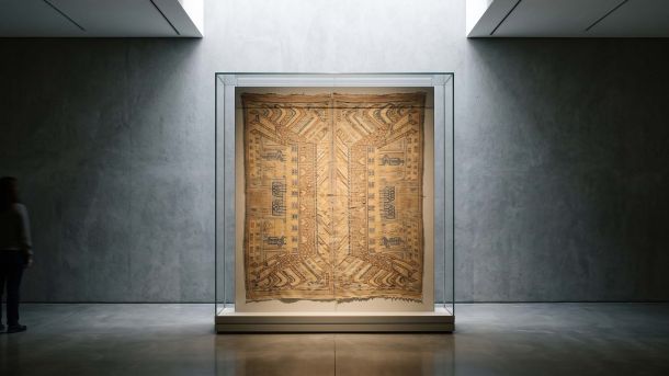 A museum vitrine holding a woven ritual textile used to screen a doorway, lit from above in a quiet gallery
