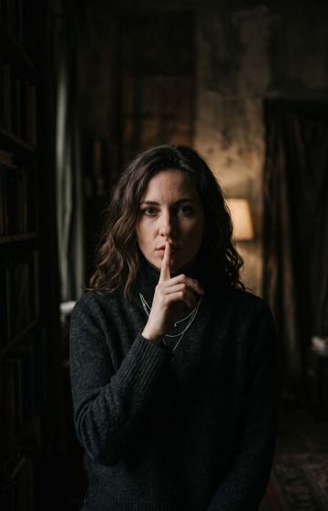 An artistic woman in dark shadows with her finger to her mouth shushing, evoking silence