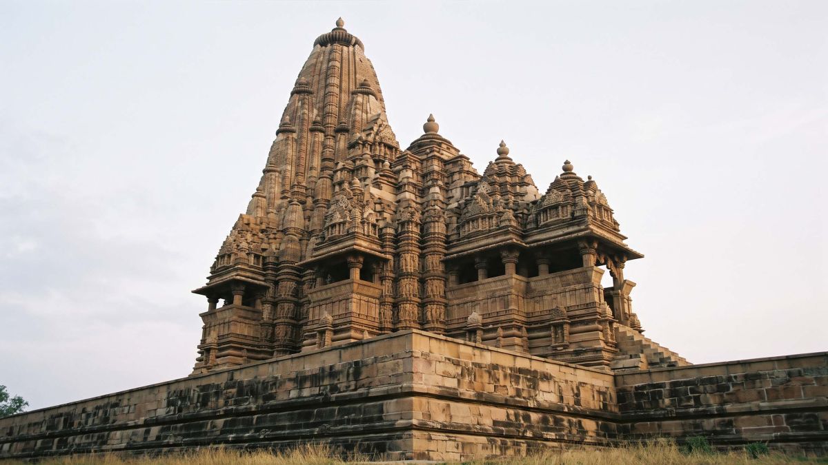 A visitor standing at the base of a carved sandstone Khajuraho temple in Madhya Pradesh at golden hour, dwarfed by its banded relief walls and tiered spire